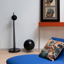 Cabasse iO3 Loudspeaker on stand in black with a Pearl Sub in black beside it.  There's a table beside it and a picture resting on the table.  In the foreground is a blue chair with a colourful cushion.