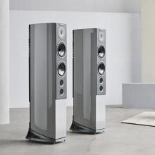A pair of tall grey speakers on a polished concrete floor in a room with white walls.