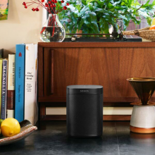 Sonos One Black next to a bowl with a lemon in with a wooden sideboard behind it with cookery books stood up to the left and a vase on top with stems of red berries in.