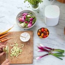 Sonos One White next to vegetables on a chopping board and tomatoes in a small bowl.  All on a marble worktop.